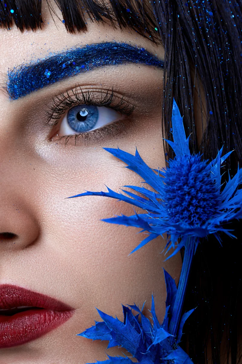 Close-up beauty portrait retouching showing a model with blue glitter eyebrows, red lips and a blue thistle flower