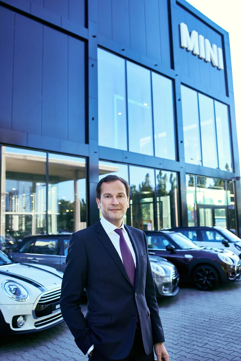Executive business portrait retouching of a manager standing outside a modern glass car showroom building